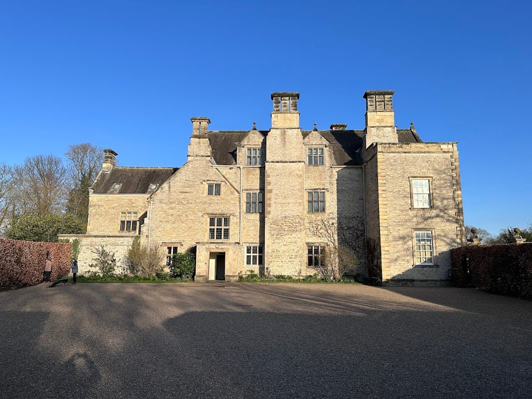 Nunnington Hall with stone facade and large windows, surrounded by greenery and a gravel car park under a clear blue sky.