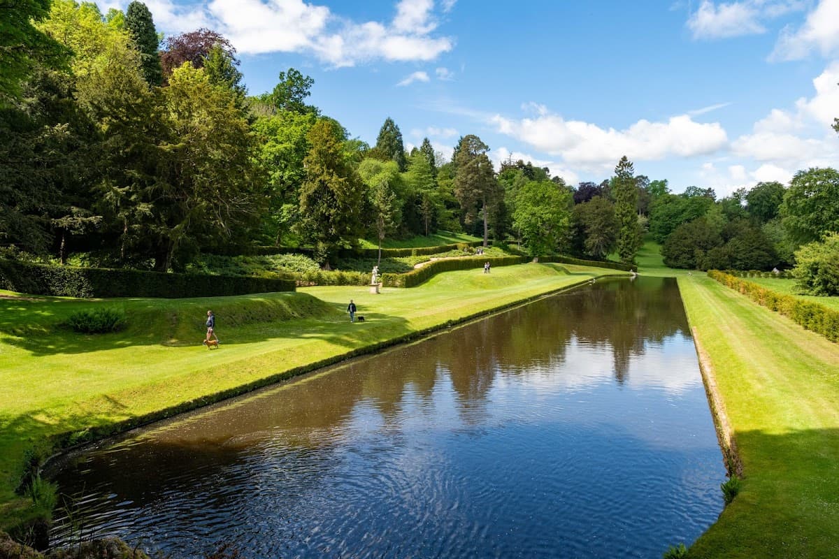 Lush green park with manicured lawns, trees, and a serene waterway under a bright blue sky.