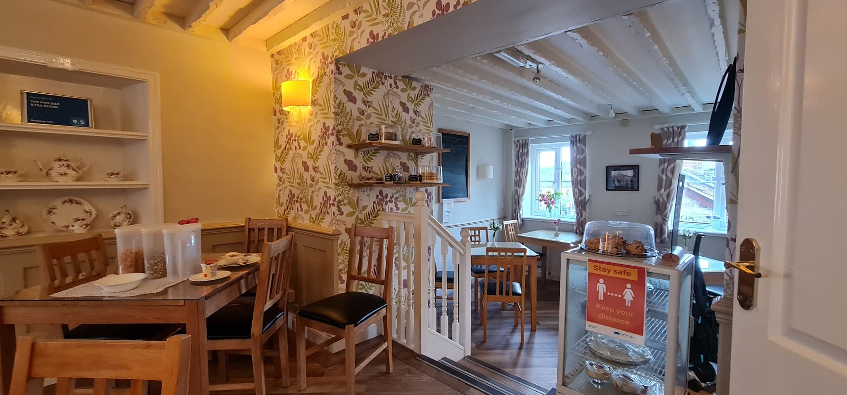 Cozy tea room with wooden furniture, floral wallpaper, and a display of baked goods near a sunny window.