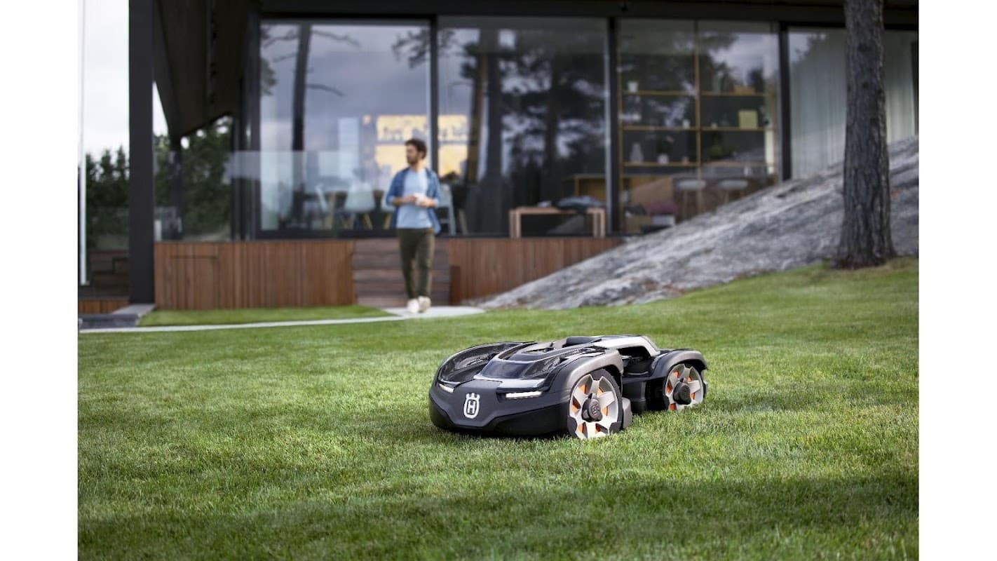 Robotic mower on green grass with a modern building and a person in the background, enjoying the outdoor space.