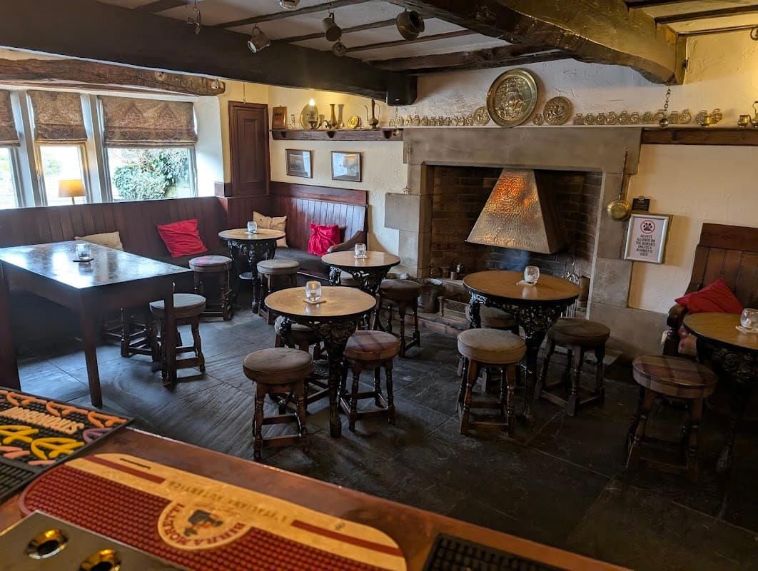 Cozy pub interior featuring wooden beams, tables, stools, and a stone fireplace with decorative elements.
