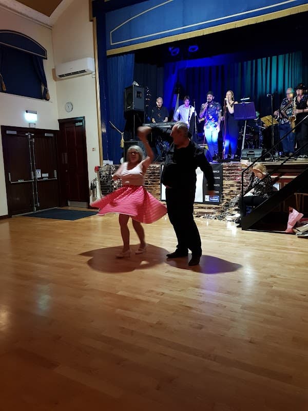 A couple dances on a wooden floor in a hall with a band performing on stage, colorful lights illuminating the scene.