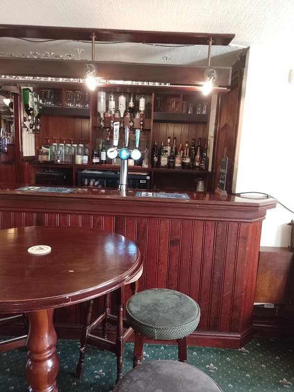 Wooden bar with beer taps, shelves of bottles behind, and a round table with a single stool in a cozy pub setting.