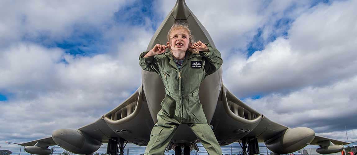 Child in a flight suit stands playfully in front of a large aircraft at the Yorkshire Air Museum.
