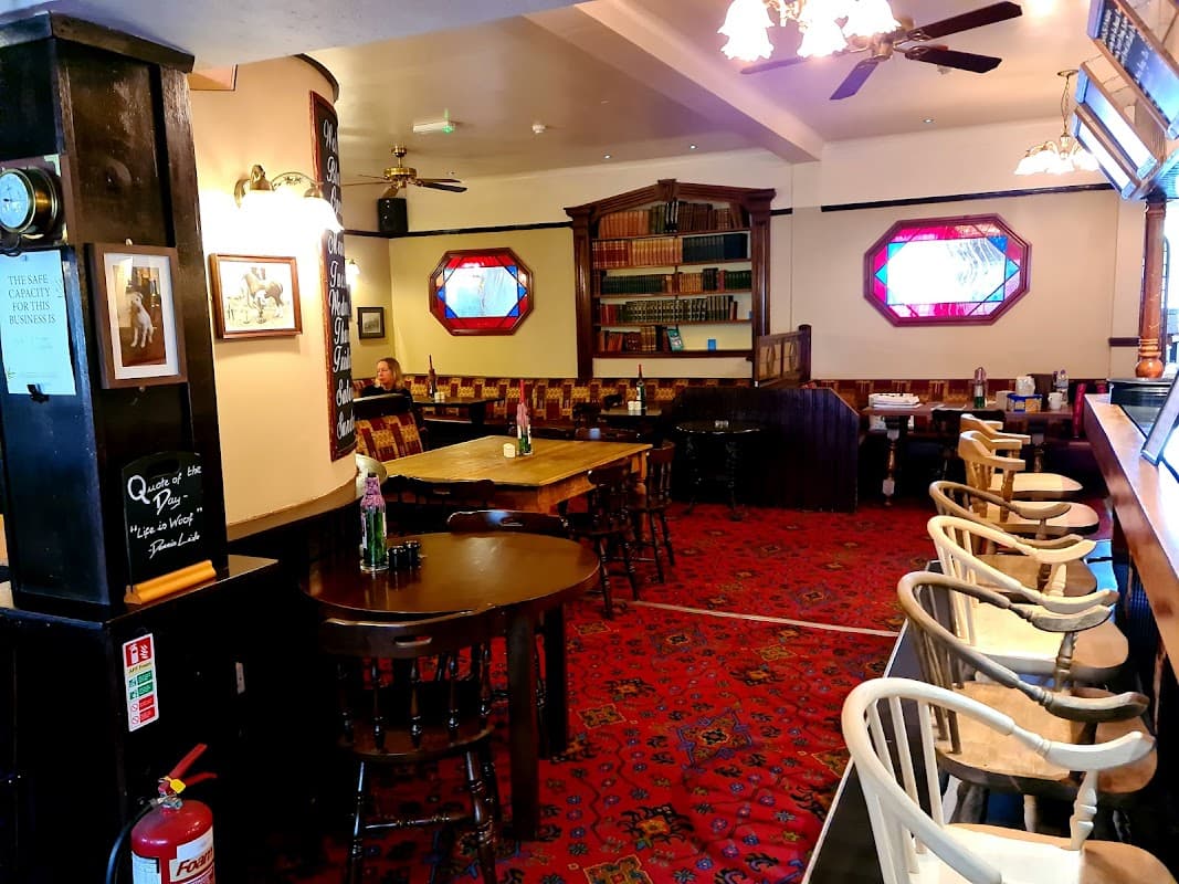 Cozy bar interior with wooden tables, chairs, patterned carpet, bookshelves, and stained glass windows.