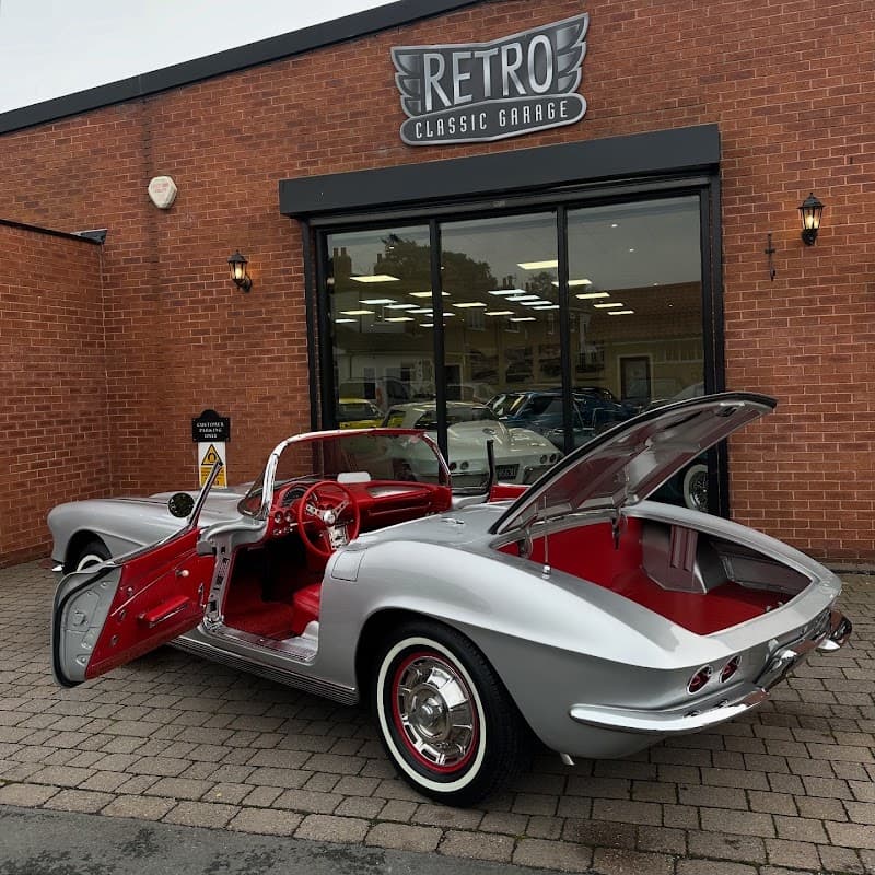Silver retro classic car with red interior parked outside a shop, featuring a sign that reads "RETRO CLASSIC GARAGE."