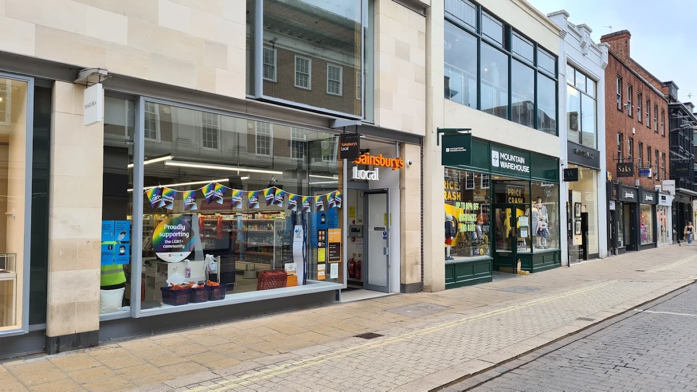 Sainsbury's Local storefront with colorful banners, surrounded by shops on a pedestrian street in Swinton, Harrogate.