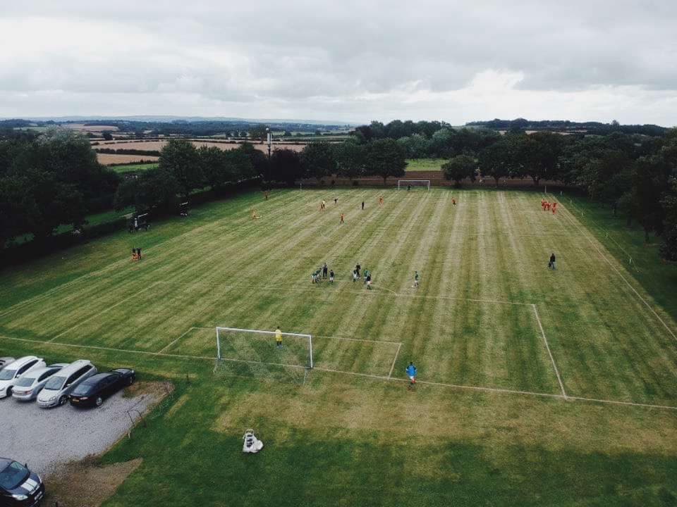 A green football pitch with players and spectators, surrounded by trees and parked cars under a cloudy sky.