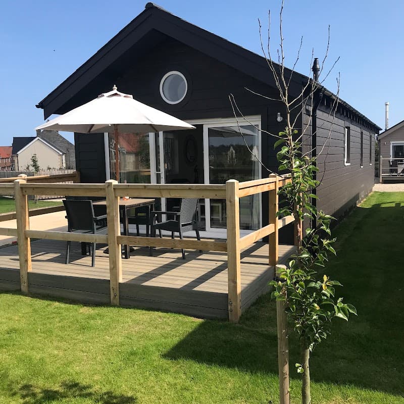 Black wooden cabin with a deck, patio furniture, and an umbrella, set in a green lawn under a clear blue sky.