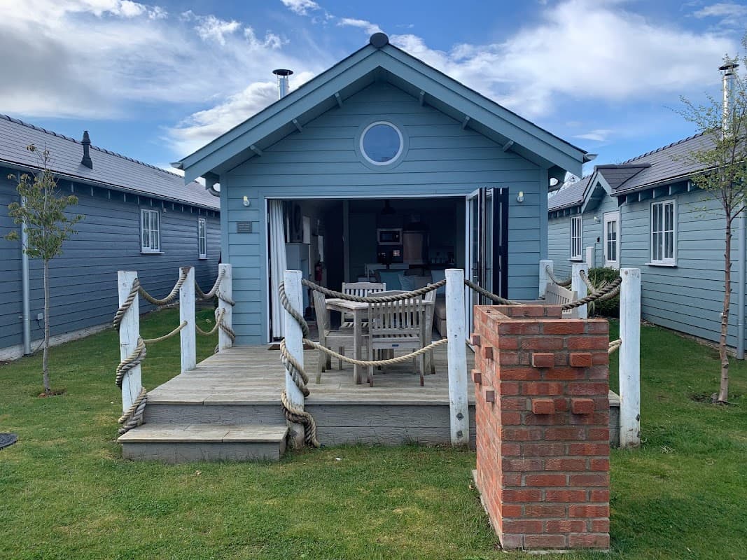 Blue beach house with a circular window, wooden deck, and brick barbecue, surrounded by green grass and other cabins.