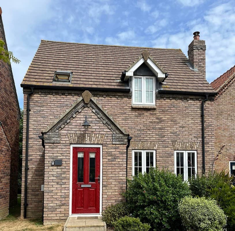 Brick cottage with a red front door, white windows, and a sloped roof, surrounded by greenery under a blue sky.