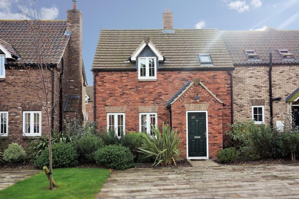 Charming brick cottage with a green door, surrounded by lush plants and a wooden pathway in a coastal setting.