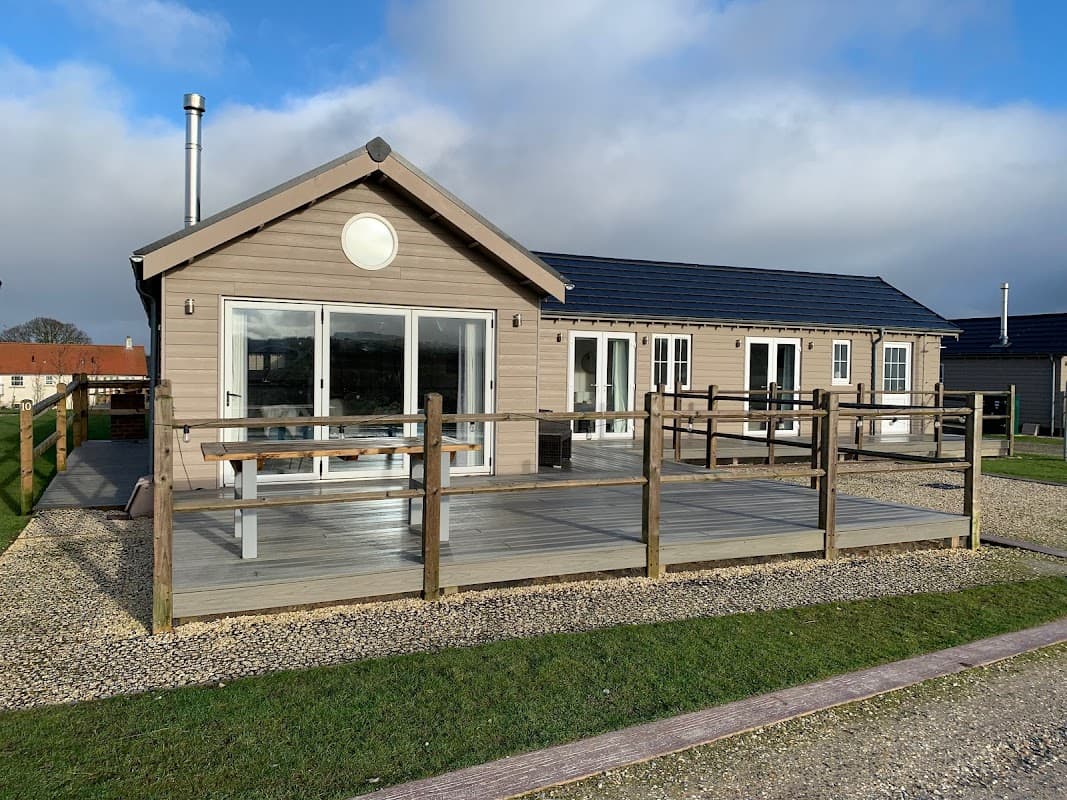 Modern lodge with large windows, wooden deck, and railings, set in a grassy area under a cloudy sky.