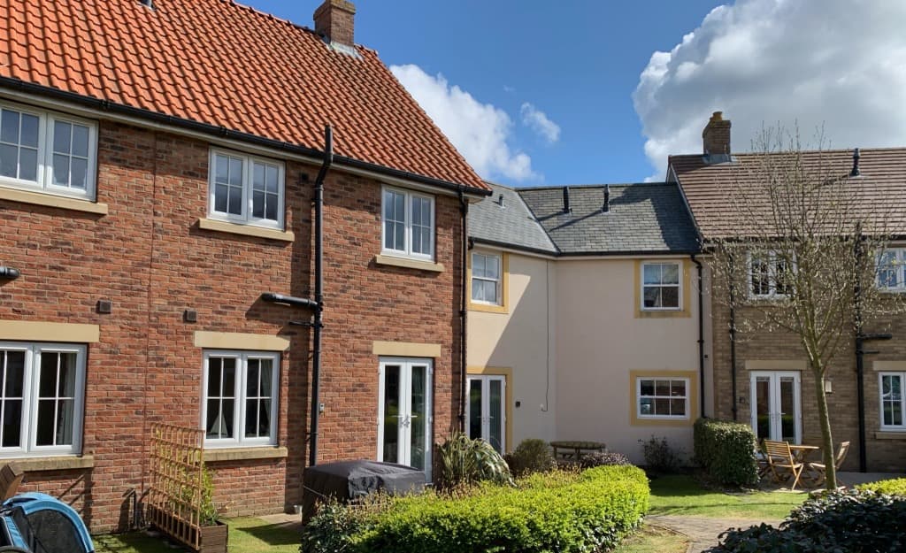 Seaside cottage with red brick and grey exterior, surrounded by green grass and trees under a partly cloudy sky.