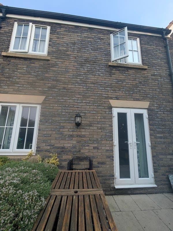 Two-story cottage with stone exterior, open window, front door, and wooden bench in a garden setting.