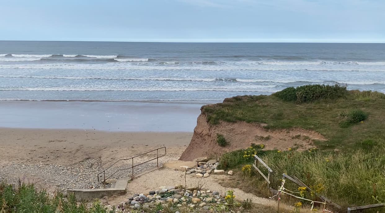 A sandy beach with gentle waves, grassy cliffs, and a rocky path leading to the shoreline under a cloudy sky.