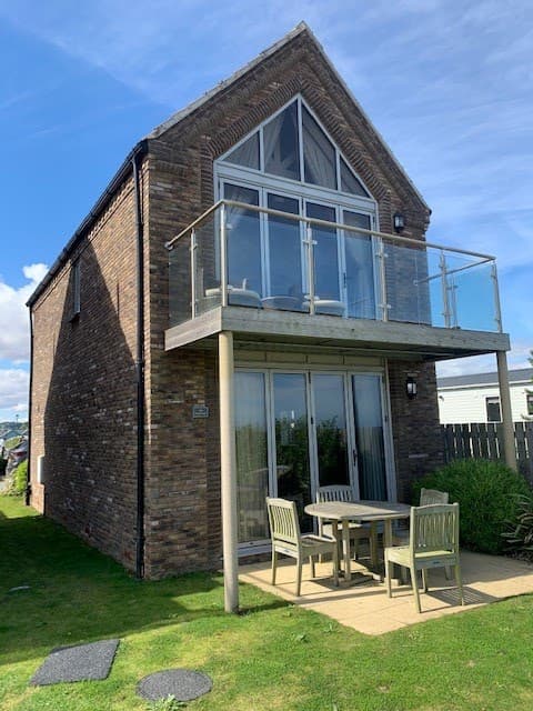 Modern brick building with large glass balcony, outdoor seating area, and green lawn under a blue sky.