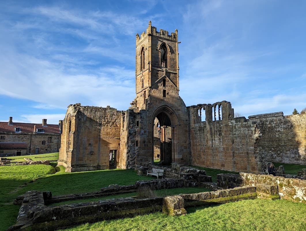 Ruins of Mount Grace Priory with a stone tower, grassy areas, and remnants of walls under a blue sky.