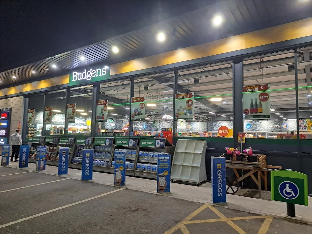 Budgens store front at night with illuminated signage, displays, and parking area. Greggs signage visible.