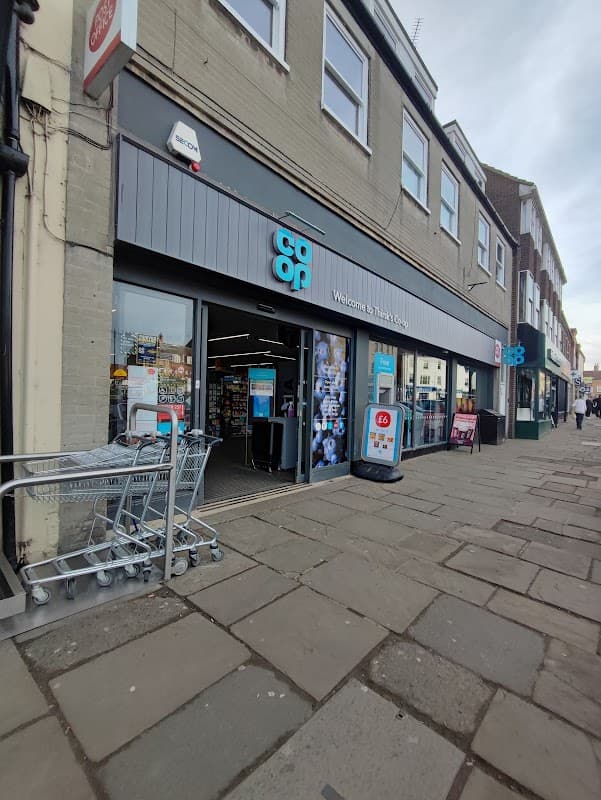 Co-op Food store entrance in Thirsk, featuring shopping carts and signage on a stone-paved market street.