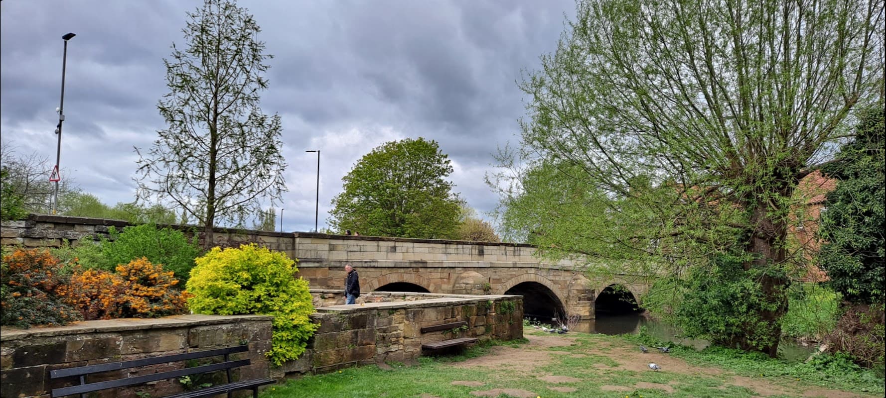 A stone bridge arches over a river, surrounded by lush greenery and colorful shrubs under a cloudy sky.