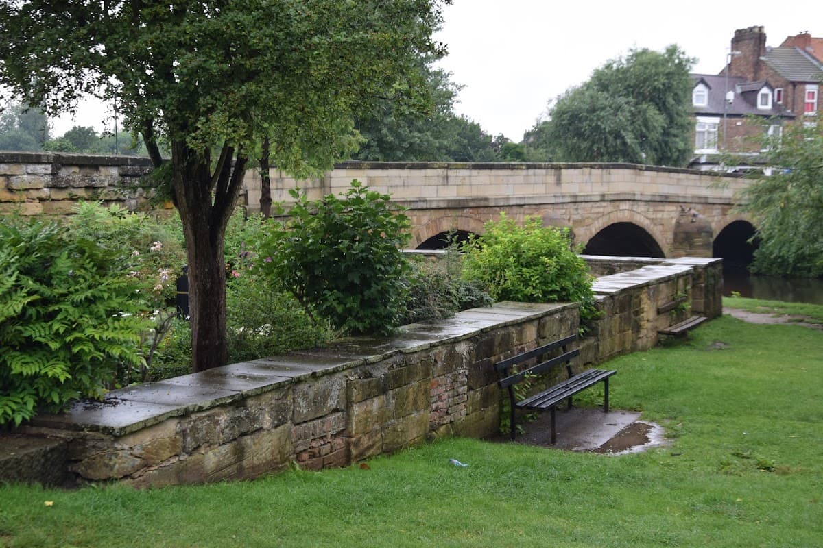 Stone wall with a bench, lush greenery, and a bridge over a river in a serene garden setting.