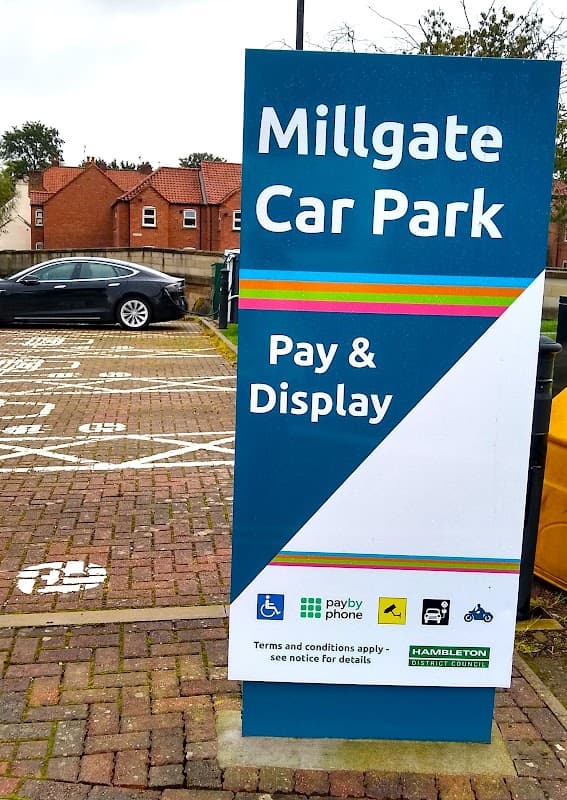 Millgate Car Park sign with "Pay & Display" instructions, featuring a car and nearby residential buildings in Thirsk.