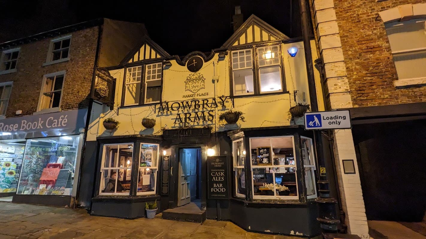 Historic bar with a half-timbered facade, illuminated at night, featuring outdoor planters and nearby shops.