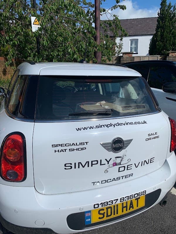 A white Mini car parked in a nursery car park with a sign for a hat shop on the rear window.