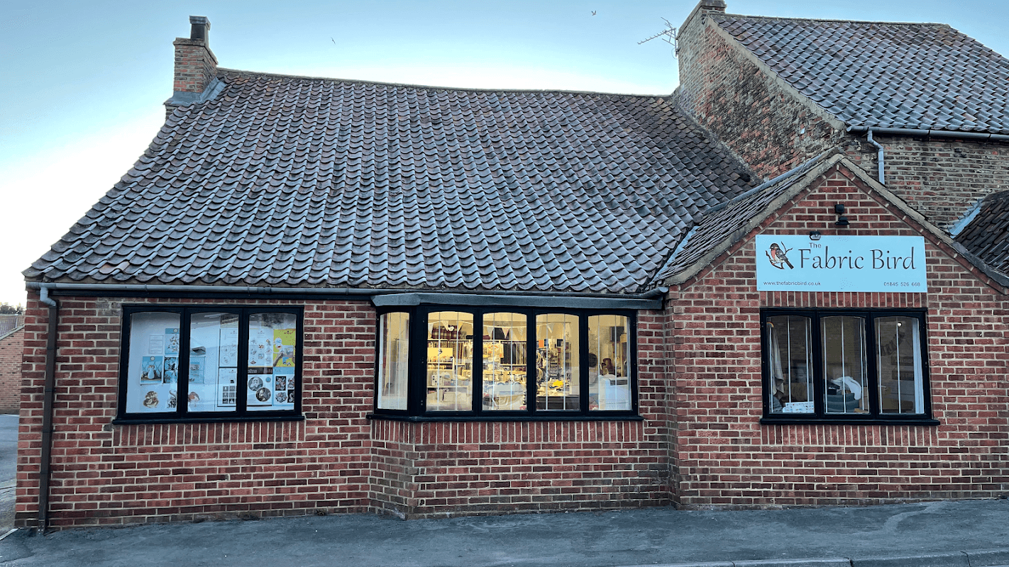 Quaint brick shop with a tiled roof, featuring large windows displaying colorful fabrics and crafts. Sign reads "Fabric Bird."