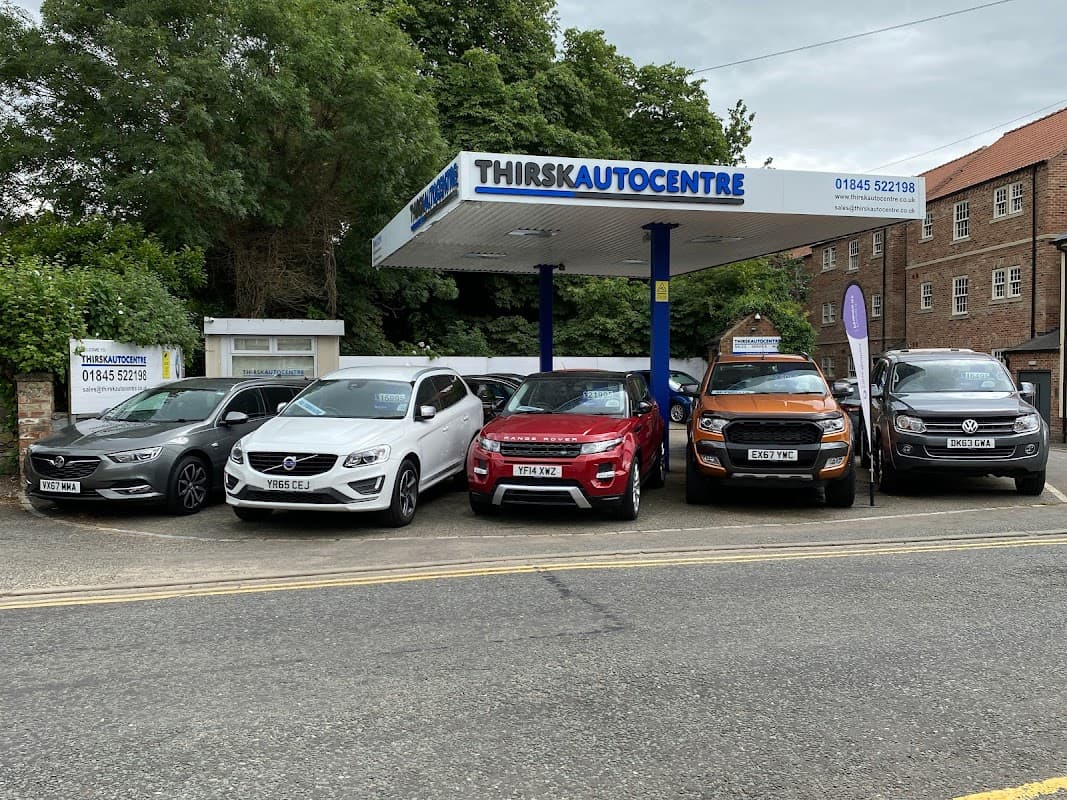 Thirsk Autocentre with a lineup of six vehicles under a canopy, surrounded by greenery and buildings in Thirsk, Yorkshire.