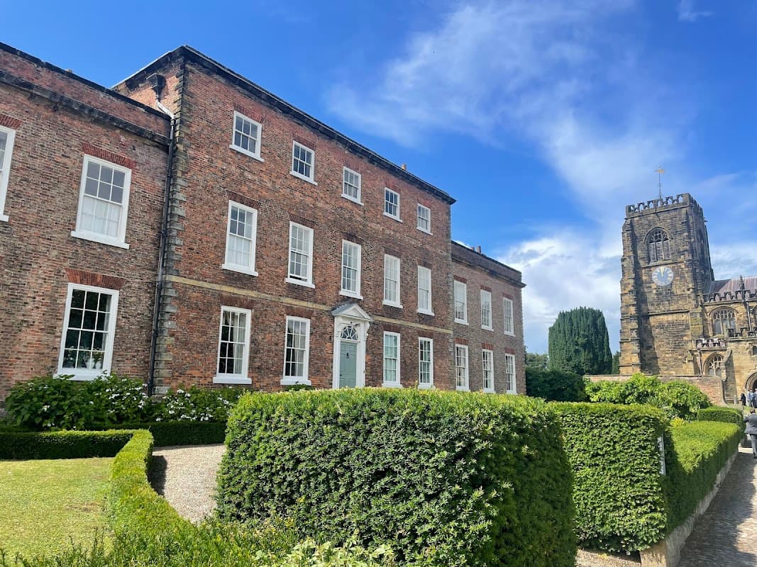 Historic brick building with large windows, manicured hedges, and a stone church tower in the background under a blue sky.