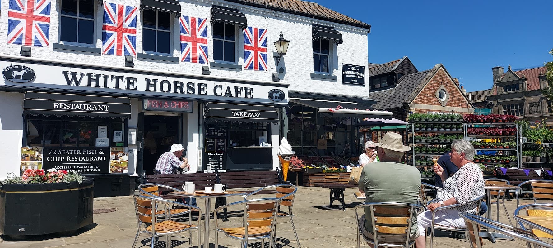 White Horse Cafe with outdoor seating, Union Jack flags, and vibrant flower displays in Thirsk Market Place.