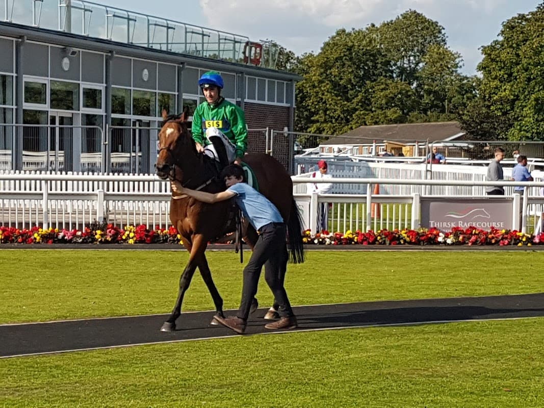 A horse with a jockey in green and blue silks is led by a person along a path at Thirsk Racecourse.