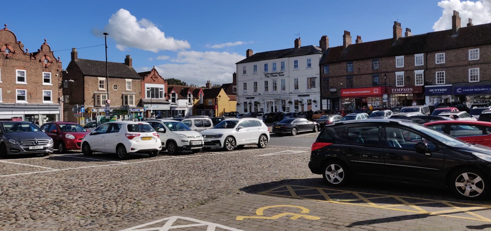 Bus stop area in Thirsk with a busy parking lot, shops, and historic buildings under a blue sky with clouds.