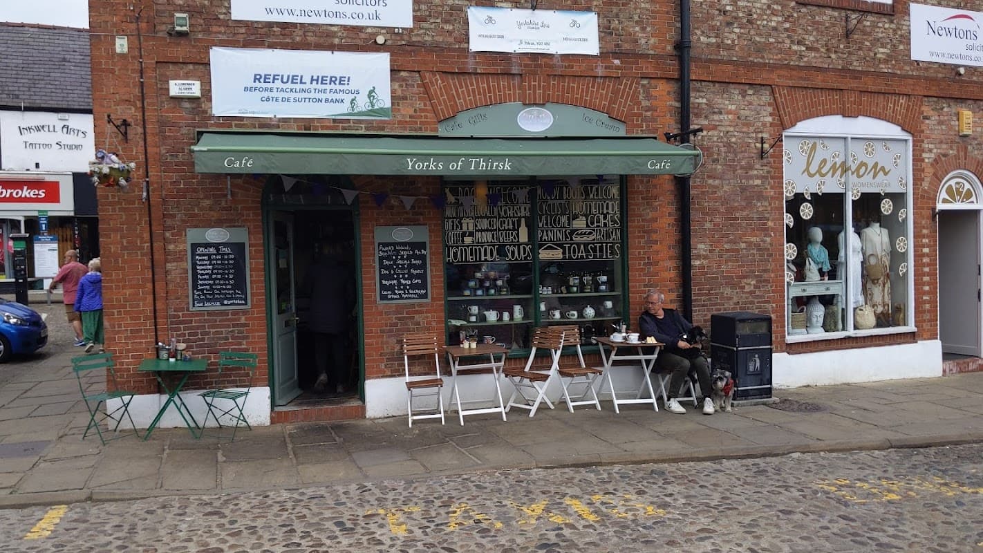 Quaint restaurant with outdoor seating, green awning, and a man sitting with a dog on a cobbled street in Thirsk.