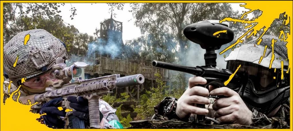 Two players in tactical gear aiming paintball guns at each other, with a wooden structure and smoke in the background.