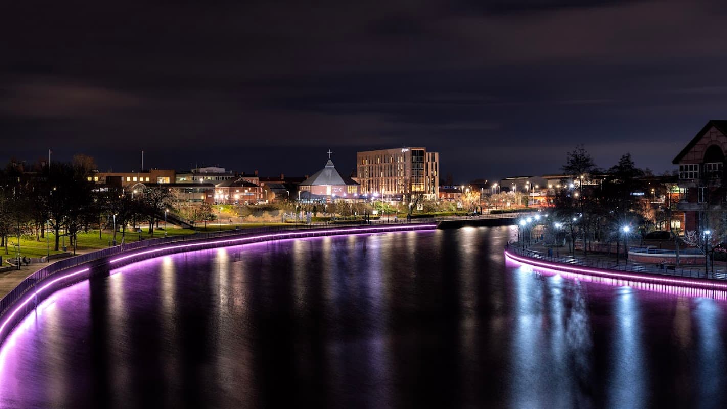 Riverside scene at night with illuminated paths, buildings, and a calm river reflecting lights in Thornaby, Yorkshire.