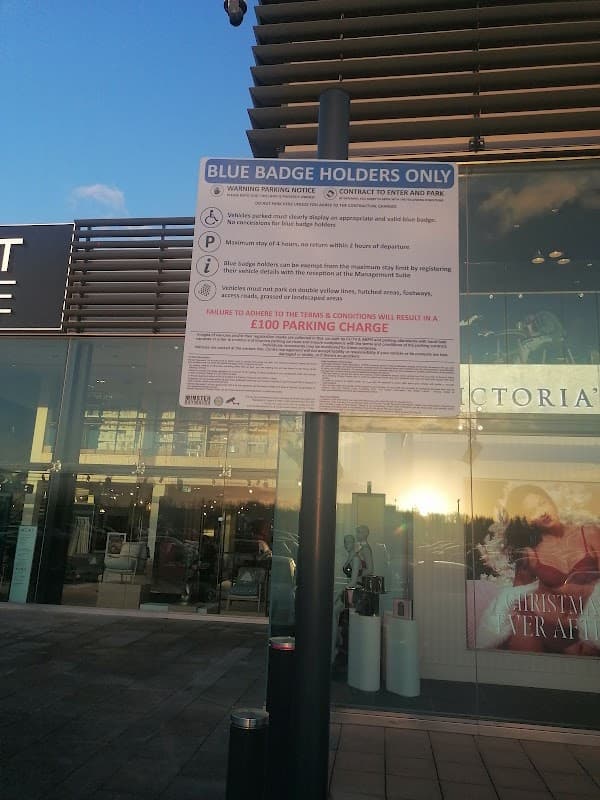 Sign for Blue Badge holders only, detailing parking rules and potential charges, with a shopping center in the background.