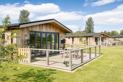Wooden lodges with large decks, surrounded by green grass and trees, under a blue sky at Cedar Retreats in Thornborough.