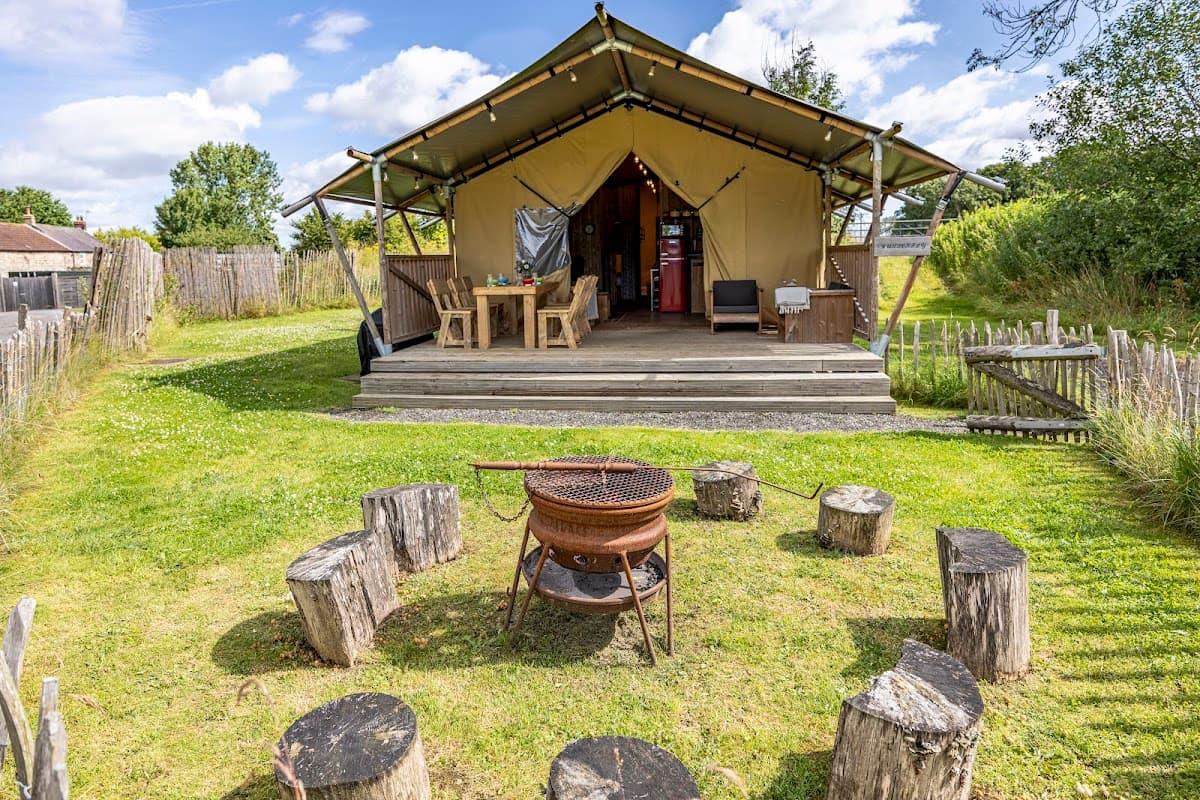 Glamping tent with wooden seating around a fire pit, surrounded by greenery and a rustic fence in Thornborough.