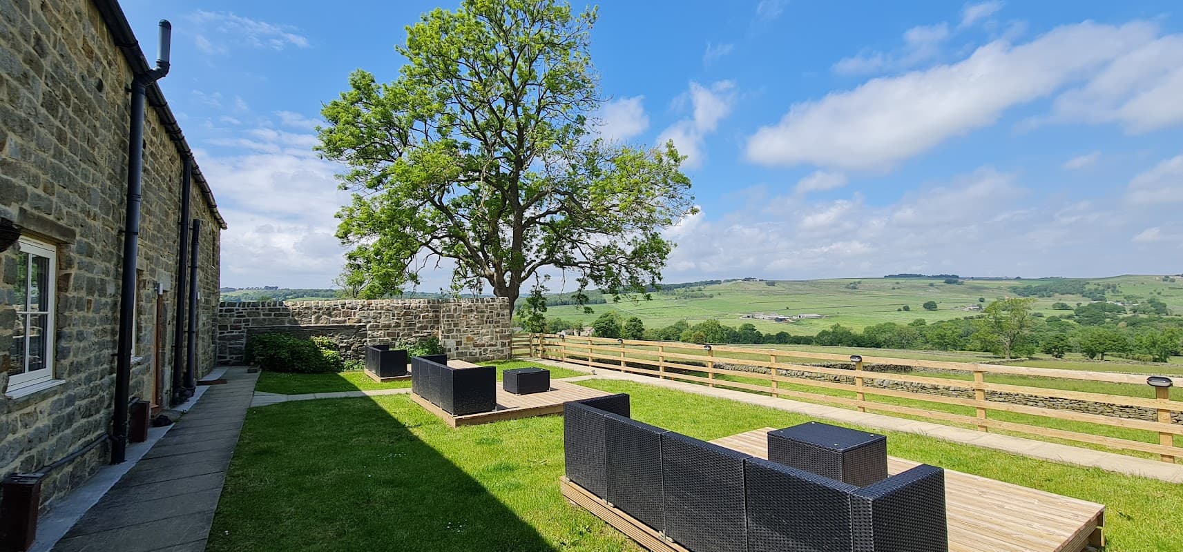 Stone cottages with outdoor seating area, lush green landscape, and a large tree under a blue sky in Thornthwaite.