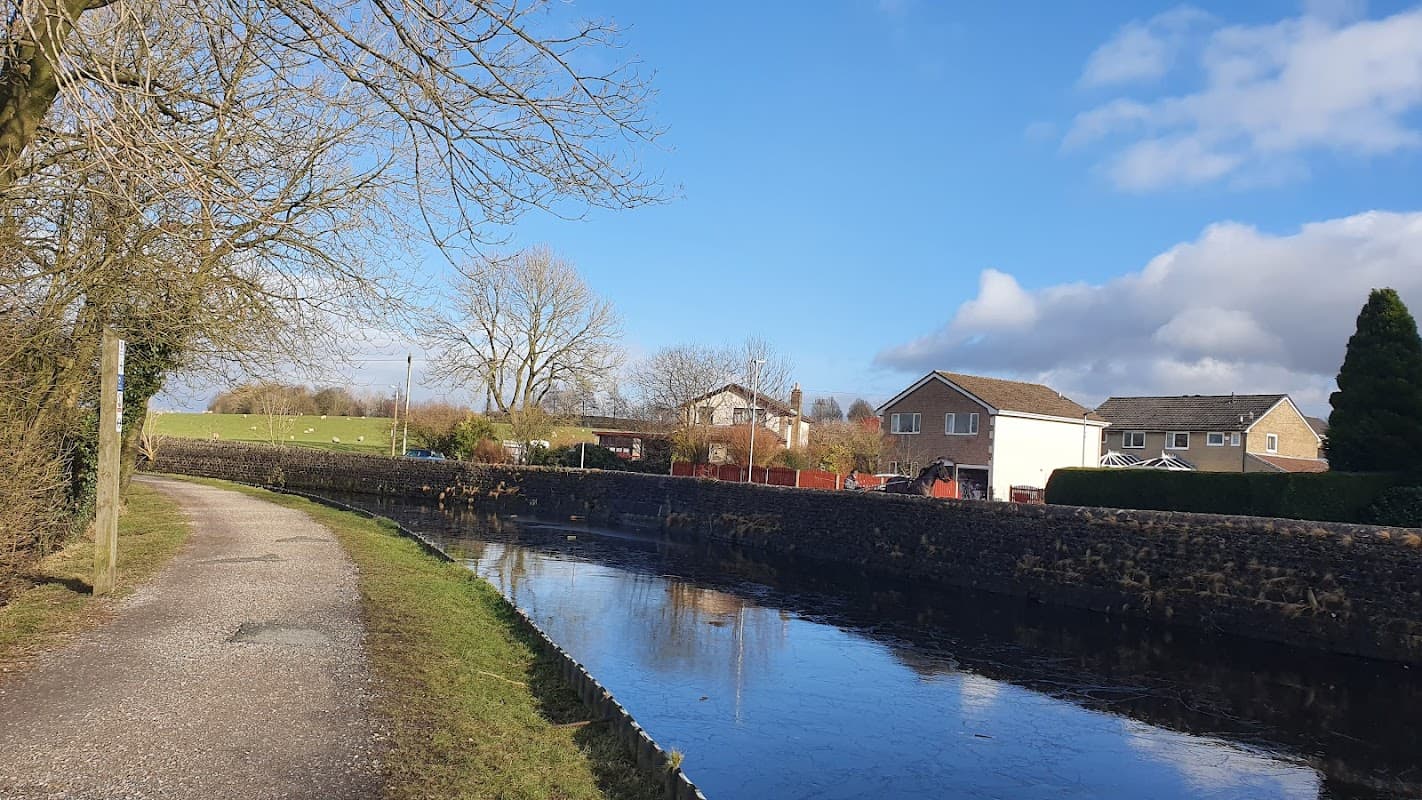 Free parking area beside a calm canal, with houses and trees under a blue sky in Thornton in Craven, Yorkshire.