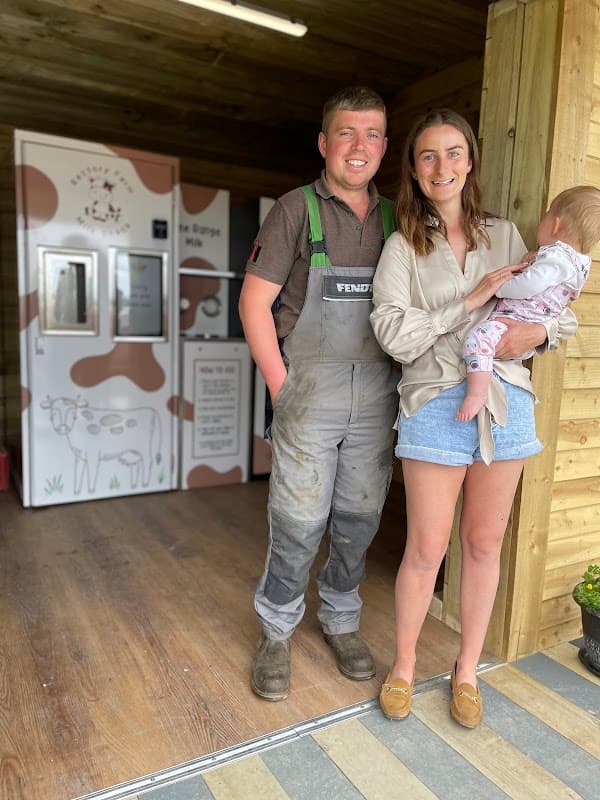 A man in overalls and a woman holding a baby stand outside a wooden shack with a milk vending machine.