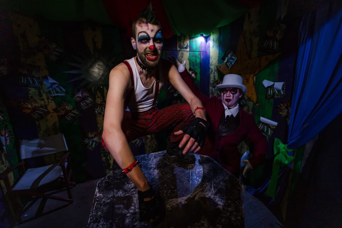 Two clowns in eerie costumes pose in a colorful, dimly lit room with theatrical decor.