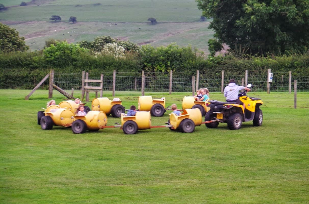 Children enjoying a ride on yellow barrel carts pulled by an ATV in a grassy field surrounded by trees and hills.