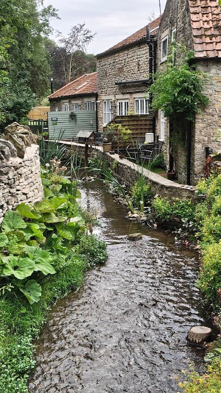 Maltongate Parking near a tranquil stream, surrounded by lush greenery and charming stone buildings in Thornton-le-Dale.