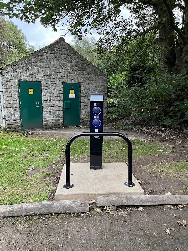 Car park with a charging station in front of a stone building with green doors, surrounded by trees and grass.