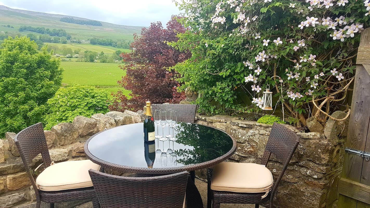 A round glass table with four wicker chairs, surrounded by greenery and flowers, overlooking a lush valley.
