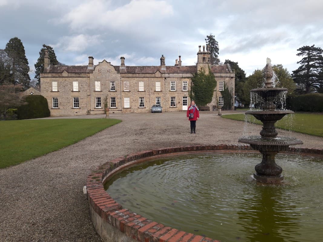 Historic Thornton Watlass Hall with a fountain in the foreground and manicured gardens, set against a cloudy sky.